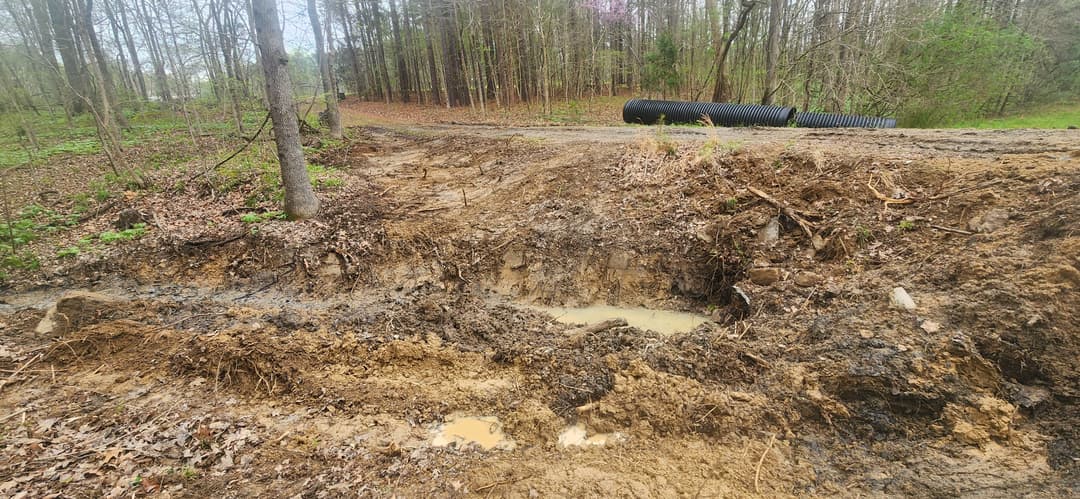 Dirt excavation site with standing water, surrounded by trees and culvert pipes nearby.