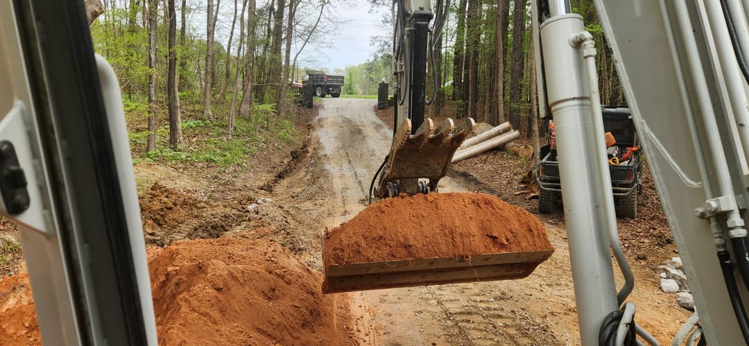 Excavator bucket lifting orange dirt on a forest road construction site.