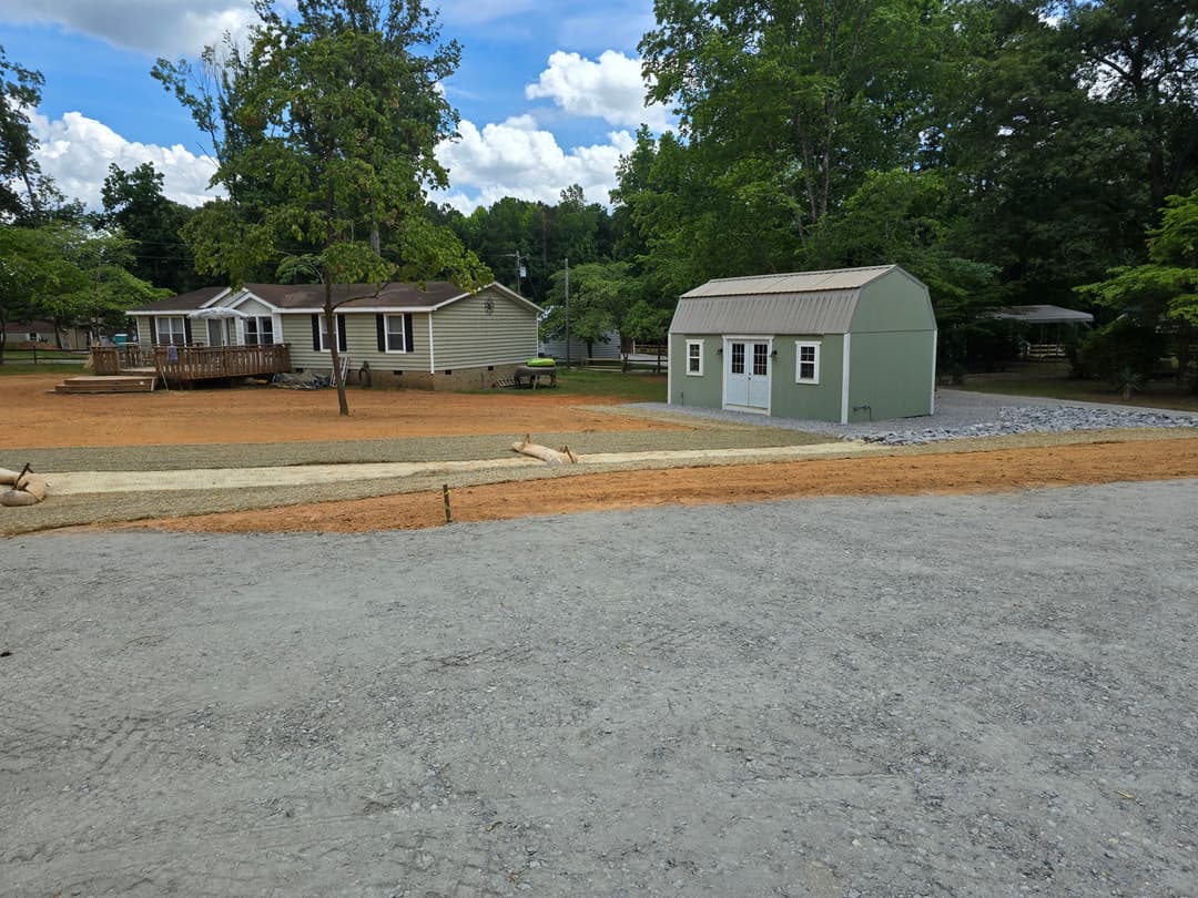 View of a backyard with a small storage shed, gravel path, and two homes under a blue sky.