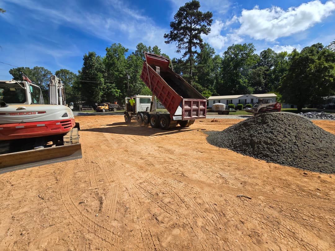 Dump truck unloading gravel at a construction site with machinery and trees in the background.