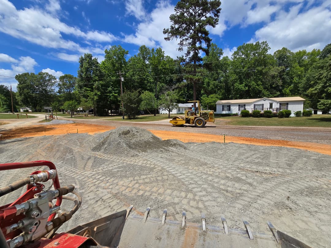 Construction site with grading machinery and gravel in a residential area under a blue sky.